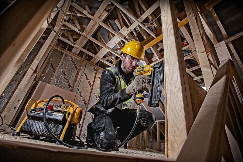 Arbeiter nutzt DEWALT Nagelpistole mit Kompressor auf einer Baustelle, trägt Schutzkleidung.