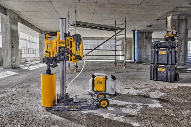 Eine DEWALT-Kernbohrmaschine mit einem gelben zylindrischen Bohraufsatz und angeschlossenem Wassertank steht auf dem Boden einer Betonbaustelle. Im Hintergrund sind Gerüste und stapelbare Aufbewahrungsboxen von DEWALT zu sehen.