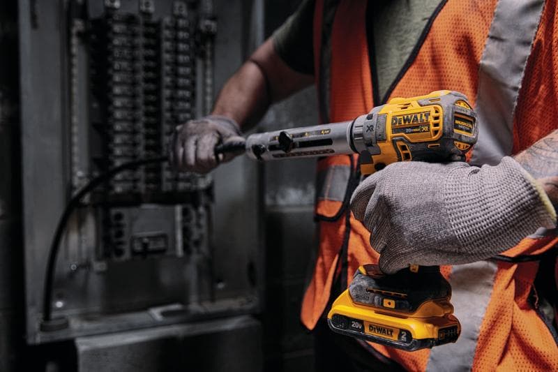 Eine Person mit Handschuhen und orangefarbener Sicherheitsweste benutzt ein kabelloses 20V DEWALT Elektrowerkzeug, um an einem elektrischen Schaltschrank zu arbeiten.