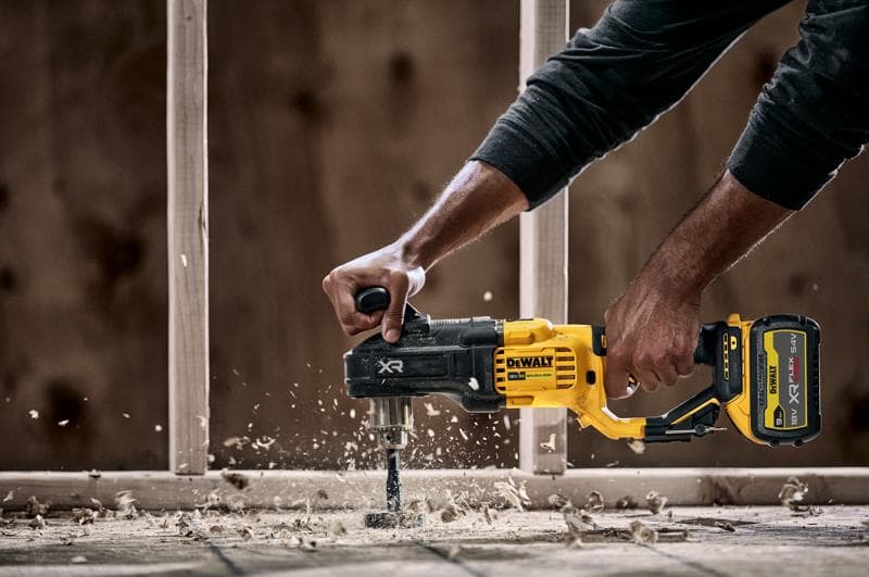 Eine Person benutzt einen DEWALT DCD444N Akku-Bohrmaschine mit Seitengriff, um durch Holz zu bohren. Dabei entstehen Holzspäne und Staub. Im Hintergrund ist eine Baukonstruktion zu sehen.