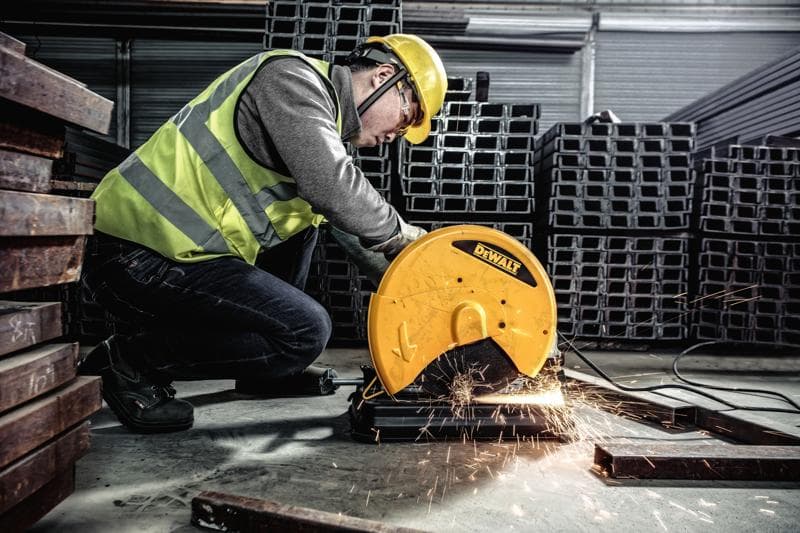 Eine Person mit Schutzkleidung bedient eine DEWALT-Metallsäge, Modell D28730, um Metallstangen in einer industriellen Umgebung zu schneiden. Funken fliegen vom Schneidbereich.