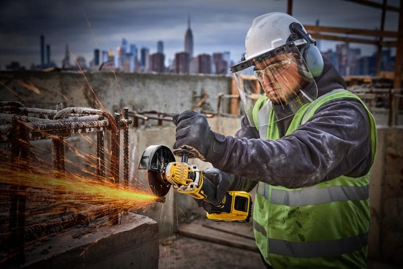 Arbeiter nutzt DEWALT DCG405P2 Winkelschleifer bei Baustelle, trägt Schutzkleidung, Stadt im Hintergrund.