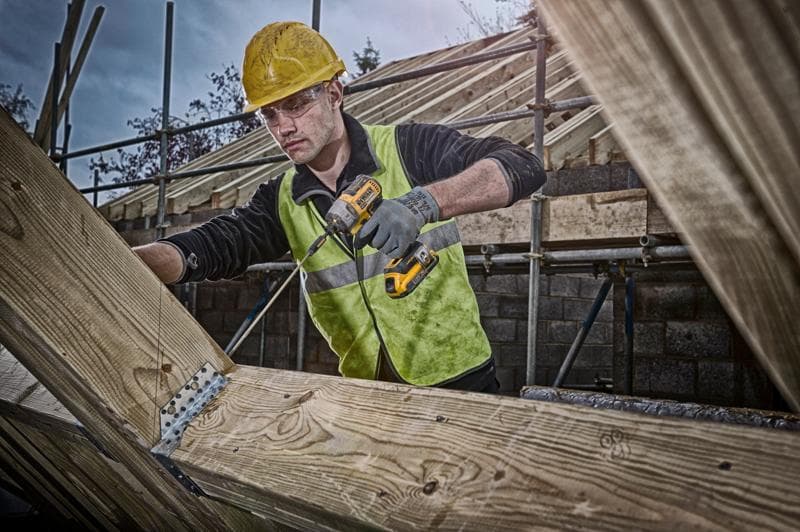 Person mit DEWALT Akkubohrer arbeitet an Holzträgern auf Baustelle, trägt Sicherheitsweste und Helm.