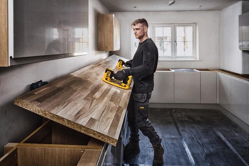 Eine Person mit verschwommenem Gesicht installiert eine Holzarbeitsplatte in einer Küche mit einem DEWALT Elektrowerkzeug. Die Küche ist modern mit hellen Schränken und einem großen Fenster im Hintergrund.