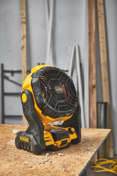 Ein tragbarer, kabelloser DEWALT Ventilator (Modell DCE512B) steht auf einer Holzwerkbank in einer Werkstatt mit Baumaterialien im Hintergrund.