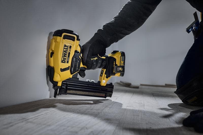 Eine Person verwendet eine gelb-schwarze DEWALT DCN210 Akku-Nagelpistole, um eine Holzleiste am unteren Rand einer Wand zu befestigen.