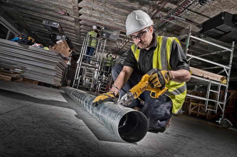 Arbeiter schneidet Rohr mit DEWALT Akku-Blechschere. Sicherheitsausrüstung und Werkzeuge sichtbar.