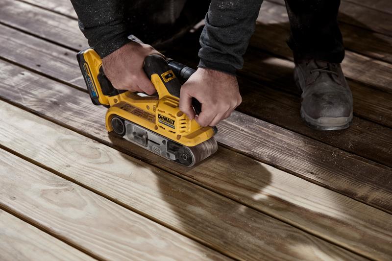 Eine Person verwendet einen gelben kabellosen DEWALT Bandschleifer auf Holzplatten, um deren Oberfläche zu glätten.