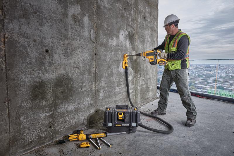 Ein Bauarbeiter mit Sicherheitsweste und Helm verwendet einen kabellosen DEWALT-Bohrer mit Staubabsaugungsaufsatz, um eine Betonwand zu durchbohren. Der Arbeiter steht auf einem großen Gebäude mit Stadtansicht im Hintergrund. Mehrere DEWALT-Werkzeuge und ein Werkzeugkoffer liegen in der Nähe auf dem Boden.