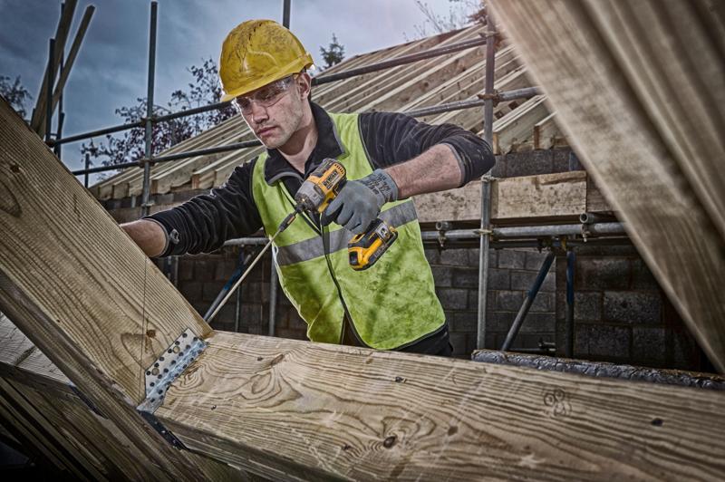 Person mit DEWALT Akkubohrer arbeitet an Holzträgern auf Baustelle, trägt Sicherheitsweste und Helm.