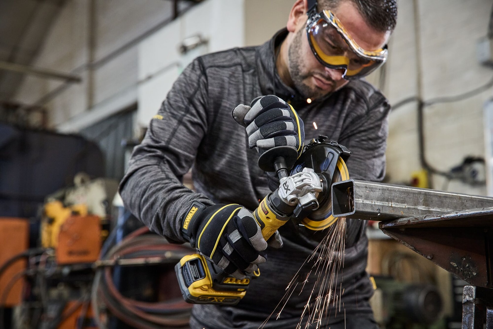 A person wearing protective gloves operates a DEWALT cordless angle grinder, model DCG404, to cut a metal bar in an industrial workshop. Sparks are flying from the grinding area.