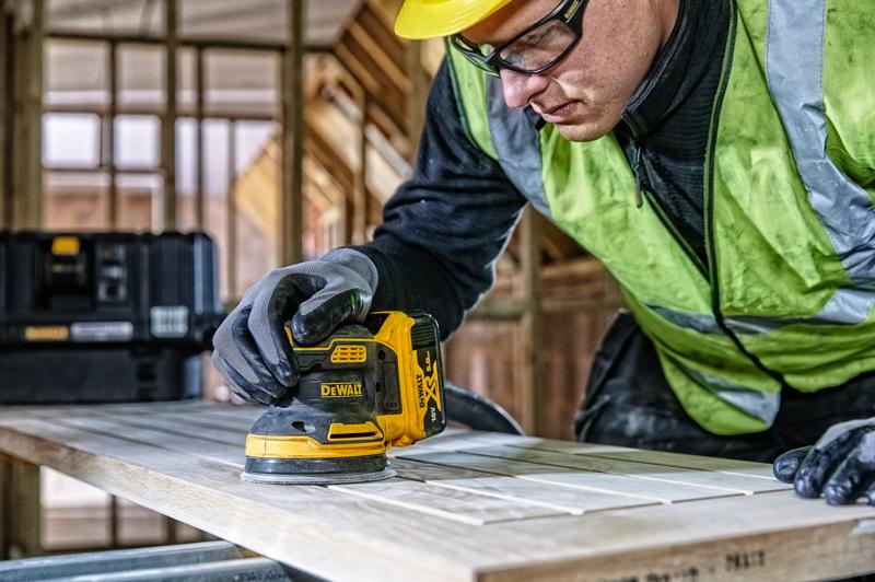 DEWALT DCW210, DCW210D1 Schleifer im Einsatz auf Holz, Baustelle, Person mit Handschuhen und Sicherheitsweste.